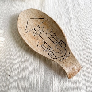 Peut inclure: Une cuillère en céramique brune avec un motif de champignons. La cuillère a une surface rugueuse et texturée et est décorée d'un contour noir de plusieurs champignons.