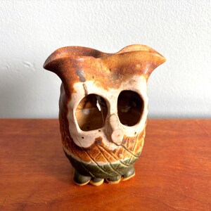 May include: A ceramic owl-shaped vessel featuring a mottled brown and tan glaze on the upper portion and a textured green base. The piece has two large, hollow eye openings, a small central beak, and a flared, wavy rim at the top opening.