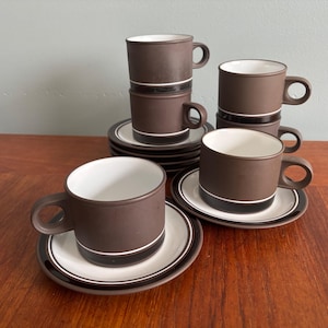 Peut inclure: Ensemble de tasses à café et soucoupes en céramique marron. Les tasses sont cylindriques avec des anses et un intérieur blanc. Les soucoupes sont blanches avec des bordures marron et noires. L'ensemble comprend plusieurs tasses et soucoupes.