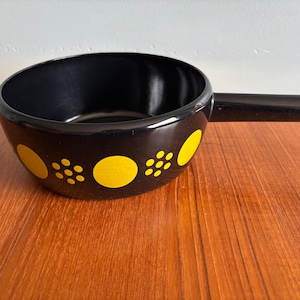 May include: A black ceramic fondue pot with a long handle. The pot has a pattern of yellow circles and clusters of yellow dots around the rim. The pot is on a wooden surface.