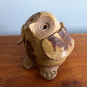 May include: A ceramic owl figurine in shades of tan and brown. The owl has large, round eyes and a small beak. The wings and body are decorated with darker brown markings. The owl is perched on two small feet.