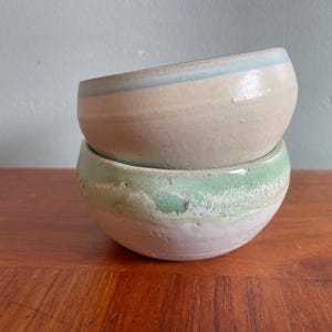 May include: Two handmade ceramic bowls stacked. The top bowl is beige with a light blue rim. The bottom bowl is green with a white base. The bowls are on a wooden surface. The bowls are likely for serving food.