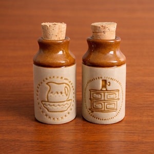 May include: Two small ceramic bottles with cork stoppers. The bottles are tan with brown tops and feature embossed designs of a jug and a cabinet. The bottles are displayed on a wooden surface.