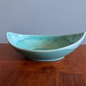 May include: A turquoise ceramic bowl, boat-shaped with a smooth, glossy finish and curved rim. The interior has a lighter turquoise shade, creating contrast. The bowl is likely suitable for serving food.