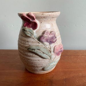 May include: A small, beige ceramic vase with a textured surface and a flared rim. The vase is adorned with three-dimensional, hand-painted flowers in shades of pink and purple, with green leaves. The vase is approximately 13 cm tall.