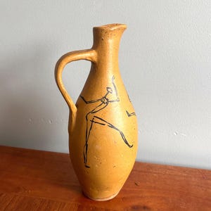 May include: A tan ceramic jug with a handle and spout. The jug is decorated with black line drawings of figures. The jug is on a wooden surface.