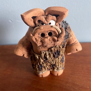 May include: A handmade ceramic figurine of a fantastical creature. The figure has a brown body, large ears, and a textured fur-like chest. The face features two large eye holes and a detailed snout. The figurine is standing on a wooden surface.
