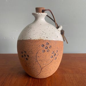 Peut inclure: Une gourde en céramique artisanale avec un bouchon en liège et une lanière en cuir. La moitié supérieure est blanche mouchetée et la moitié inférieure est marron texturé avec un motif floral gravé. La gourde mesure environ 18 cm de haut.