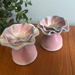 Peut inclure: Deux pièces décoratives en céramique. Chacune a une base rose arrondie soutenant un dessus froissé en forme de fleur. Les dessus présentent des nuances de rose, crème et bleu. Les pièces sont exposées sur une surface en bois.