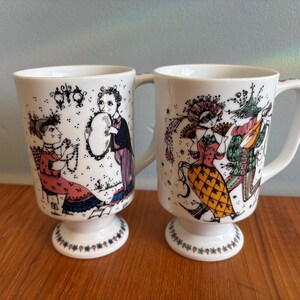 May include: Two white ceramic mugs with pedestal bases and handles. Each mug features a colorful illustration of figures in period clothing. The mugs have a decorative floral pattern around the base and are set on a wooden surface.