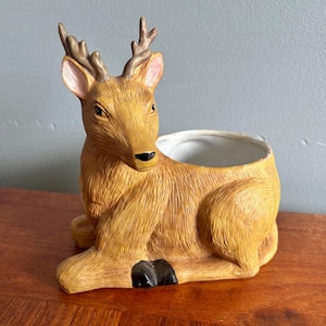 May include: A ceramic planter shaped like a resting deer with textured golden-brown fur, light pink inner ears, dark brown antlers, and black hooves. The hollow back of the deer serves as a container with a smooth white interior finish.