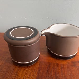 May include: A brown ceramic creamer and sugar bowl set. The sugar bowl has a lid with a brown, black, and white design. The creamer has a handle and spout. Both pieces have a white and black stripe near the base.