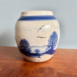 May include: A speckled cream-coloured ceramic vase with a rounded body and a wide opening. The vase features a blue painted landscape design depicting trees, rolling hills, and birds in flight, with a solid blue band encircling the upper rim.