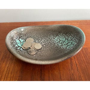 May include: A small, oval-shaped ceramic dish with a speckled, dark grey and turquoise glaze. The dish has a rough, textured surface and is filled with several silver coins.