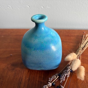 Peut inclure: Un petit vase en céramique arrondi avec un vernis bleu brillant et varié, passant d'un turquoise clair au niveau du col à un bleu plus profond et tacheté à la base. Le vase présente une ouverture circulaire étroite et un corps bombé lisse.
