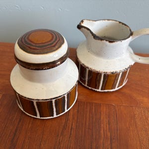 May include: A ceramic lidded sugar bowl and a matching creamer pitcher. Both pieces feature a speckled off-white glaze with a dark brown and tan vertical striped pattern around the base. The lid has a circular brown and tan ring design.