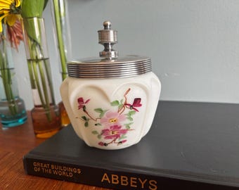 May include: A white, vintage jar with a silver lid and a floral design. The jar has a rounded shape with a series of vertical indentations. The floral design features pink and red flowers with green leaves. The jar is sitting on a dark book.