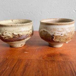 Peut inclure: Deux bols en céramique faits à la main avec un aspect rustique et texturé. Les bols ont un extérieur beige clair avec une bande marron plus foncée au milieu. L'intérieur est de couleur beige clair. Les bols sont sur une surface en bois.