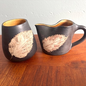 May include: A ceramic creamer and a matching cup featuring a matte black exterior with a textured, light-colored leaf imprint on the front. Both pieces have a smooth, warm yellow interior glaze and a rounded, organic shape with a curved handle.