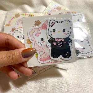 May include: A collection of stickers featuring cartoon characters. One sticker shows a white cat in a black suit with a pink tie. Another sticker shows a pink cat in a pink dress. Other stickers are visible in the background.