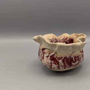 May include: A small, white ceramic bowl with a red and white glaze. The bowl has a unique, organic shape with a ruffled edge.