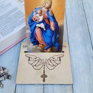 May include: A wooden stand with a cutout of two hands holding a rosary. The stand has a slot to hold a religious card featuring a painting of the Virgin Mary holding the baby Jesus. The card has the text "Vobis" at the bottom.