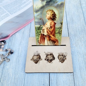 May include: A wooden card holder with a vintage religious image of a child holding a lamb. The holder features three heart illustrations. A rosary and a book are visible in the background.