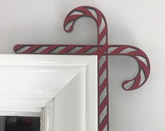 May include: Red and white candy cane shaped wooden doorstop. The doorstop is in the shape of two candy canes that are crossed at the top. The doorstop is attached to a white door frame.