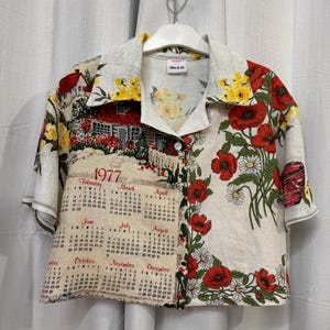 May include: A short-sleeved shirt with a unique design. One side features a calendar from 1977, while the other showcases red poppies and white daisies. The shirt also has a house and floral pattern with yellow flowers.