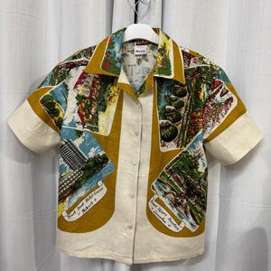 May include: Short-sleeved shirt with a cream-coloured body and a colourful, vintage-style print. The print features landscape scenes and text, with mustard-yellow accents on the collar and sleeves. The shirt is hanging on a white hanger.