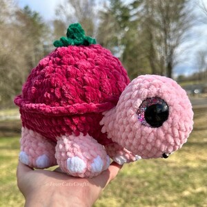 Pode incluir: Uma tartaruga de pelúcia feita à mão com uma carapaça rosa vibrante e um detalhe de folha verde no topo. A cabeça e as patas da tartaruga são rosa claro, com detalhes brancos nas patas. Os olhos grandes e brilhantes adicionam um toque lúdico a este brinquedo.