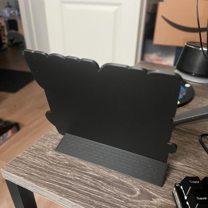 Puede incluir: Un letrero decorativo negro con una forma irregular &uacute;nica, montado sobre una base rectangular. El letrero est&aacute; hecho de un material s&oacute;lido y opaco y se apoya sobre una superficie de madera. El dise&ntilde;o es simple y moderno.
