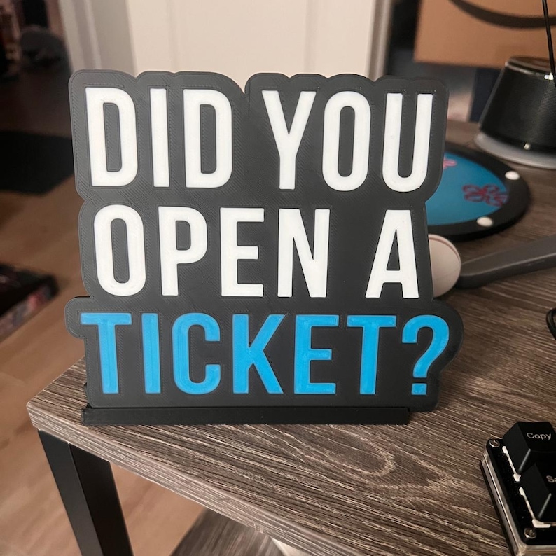 Puede incluir: Un letrero en blanco y negro con el texto "DID YOU OPEN A TICKET?" en letras blancas y azules. El letrero est&aacute; hecho de material en capas y se encuentra sobre una superficie de madera.
