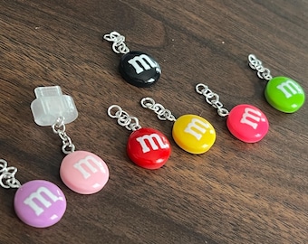 Puede incluir: Una colección de coloridos colgantes que se asemejan a caramelos, cada uno con una "m" blanca sobre un fondo de color. Los colgantes están unidos a cadenas de color plateado. Los colores incluyen rosa, morado, rojo, amarillo, verde y negro.