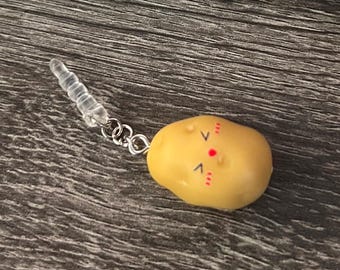 Puede incluir: Un amuleto amarillo con forma de patata con una cara sonriente, unido a una cadena plateada y un enchufe transparente. El amuleto tiene detalles rojos y negros. El fondo es una veta de madera oscura.