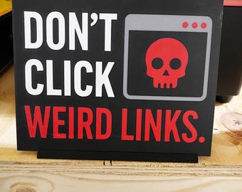 Puede incluir: Cartel rectangular negro con texto blanco que dice "DON'T CLICK WEIRD LINKS". Un icono de calavera roja está dentro de un gráfico de ventana gris. El cartel está sobre una estantería de madera.