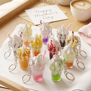 May include: A collection of colorful ice cream sundae keychains with silver keyrings. Each keychain features a different flavor, including orange, pink, green, and purple. A card that says "Happy Mother's Day" is in the background.