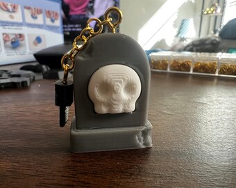 Puede incluir: Un llavero gris impreso en 3D con un diseño de calavera blanca. El llavero tiene una cadena dorada y un anillo de llave negro.