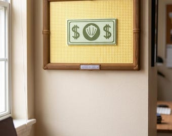 Puede incluir: Un tablero perforado rectangular de pared con un marco marrón estilo bambú y una superficie de rejilla amarilla. Centrado en el tablero hay un letrero verde rectangular con dos signos de dólar y un icono de concha. Debajo hay una etiqueta.