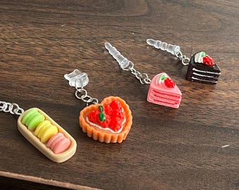 Puede incluir: Cuatro pendientes de charms con diferentes diseños de postres. Los charms están hechos de plástico e incluyen una tarta de fresa en forma de corazón, una rebanada de pastel rosa, una rebanada de pastel de chocolate y una pila de macarons.