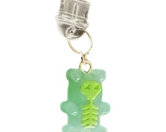 Puede incluir: Un charm plateado con un anillo dorado y un colgante de osito de goma verde. El osito de goma tiene un diseño de esqueleto alienígena verde en el centro. El charm está sujeto a un cierre plateado.