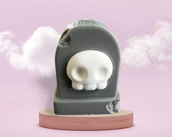 Puede incluir: Una lápida de cerámica gris con una cara de calavera blanca en la parte delantera. La lápida está sentada sobre una base de madera.
