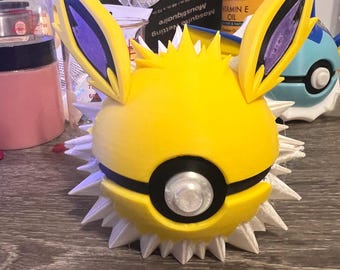Puede incluir: Una figura de Pokémon impresa en 3D de color amarillo y blanco que representa a un Pichu de orejas puntiagudas. La figura tiene un diseño de Poké Ball negro y plateado en su cuerpo. La figura tiene una base blanca y puntiaguda.