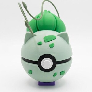 Puede incluir: Una Pokéball con temática de Bulbasaur impresa en 3D. La esfera verde claro tiene detalles en blanco y negro, con manchas verdes y un bulbo verde en la parte superior. Se apoya sobre una pequeña base morada.