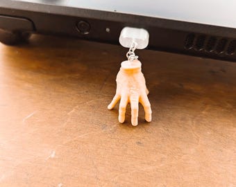 Puede incluir: Una consola de juegos portátil negra con un pequeño amuleto de mano de color canela colgando de un lado. El amuleto está unido a una pieza rectangular transparente. La consola está sobre una superficie de madera marrón.
