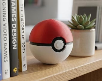 Puede incluir: Una réplica roja y blanca de Poké Ball, una esfera con una banda negra y un botón central. El objeto coleccionable está sobre una estantería de madera junto a libros como "Games Design" y una pequeña suculenta en maceta. El tema general es el juego.