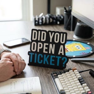 Puede incluir: Un letrero en blanco y negro con el texto "DID YOU OPEN A TICKET?" sobre un escritorio de madera. Un teclado mec&aacute;nico, un rat&oacute;n, un bloc de notas y un smartphone tambi&eacute;n est&aacute;n en el escritorio.