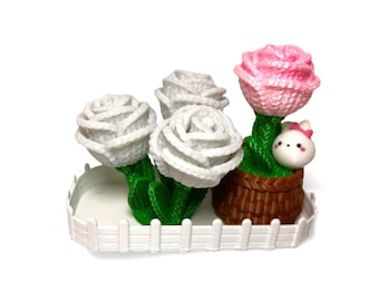 Puede incluir: Una escena de jardín en miniatura decorativa con cuatro rosas de ganchillo en una valla blanca. Tres rosas son blancas y una es rosa. Una pequeña figura de conejito está metida en una cesta marrón con la rosa rosa.