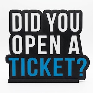 Puede incluir: Un letrero negro con el texto "DID YOU OPEN A TICKET?" en blanco y azul. El letrero es rectangular con esquinas redondeadas y se asienta sobre una peque&ntilde;a base negra. El texto est&aacute; en negrita, sin serifa.