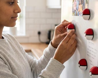 Puede incluir: Imanes redondos rojos, blancos y negros que se asemejan a una Pokeball se colocan en la puerta blanca de un refrigerador. Una persona está colocando un imán. Una foto familiar y una nota manuscrita también están adheridas al refrigerador.